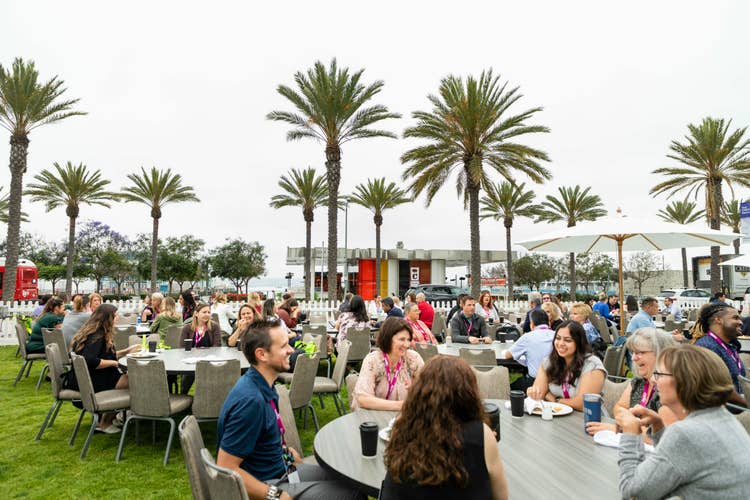 People seated at round tables outdoors, eating and talking under palm trees on a cloudy day, enjoy a relaxed atmosphere as EHS safety measures ensure everyone’s well-being.