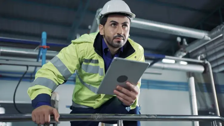 A man wearing a white hard hat and a high-visibility jacket holds a tablet while inspecting industrial equipment.