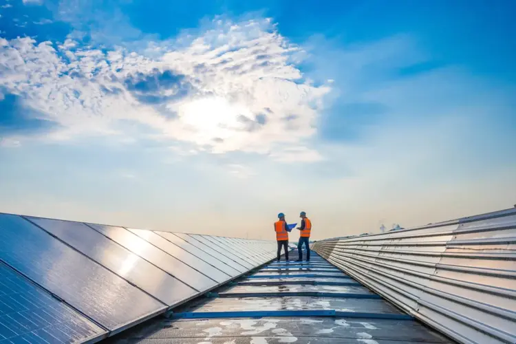 Deux travailleurs en gilets orange et casques se tiennent sur un toit à côté de panneaux solaires, discutant sous un ciel bleu vif parsemé de nuages et de soleil.
