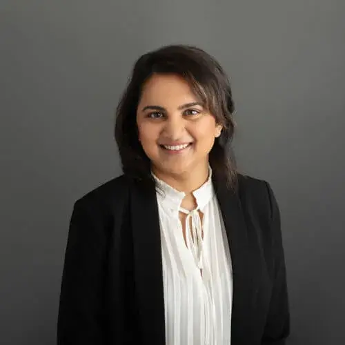 A woman with dark hair wearing a black blazer and a white blouse smiles in front of a plain gray background.