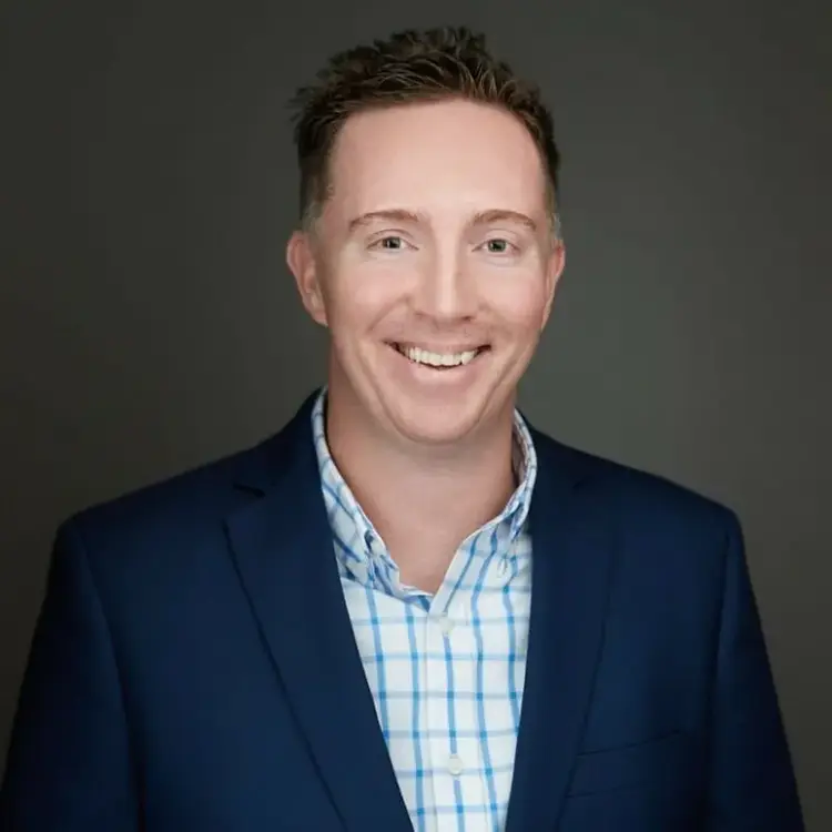 A man with short brown hair wearing a navy blue blazer and a blue-and-white checkered shirt smiles in front of a plain dark gray background.