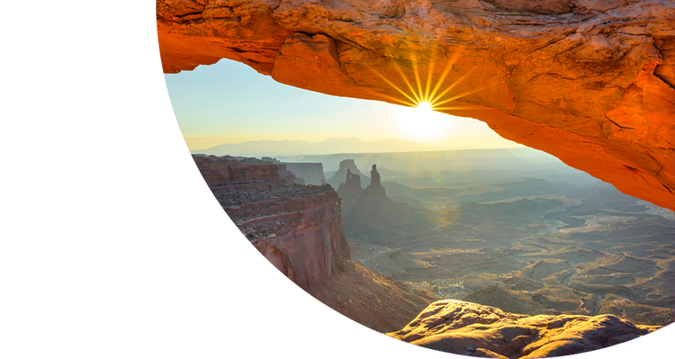 Sunrise shining through a natural red rock arch, overlooking a vast canyon landscape with rugged cliffs, mesas, and hints of Greenstone glowing in the morning light.