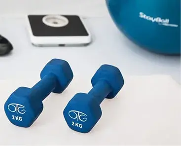 Two blue 2 kg dumbbells sit on a white surface, with a blurred scale and blue exercise ball in the background.