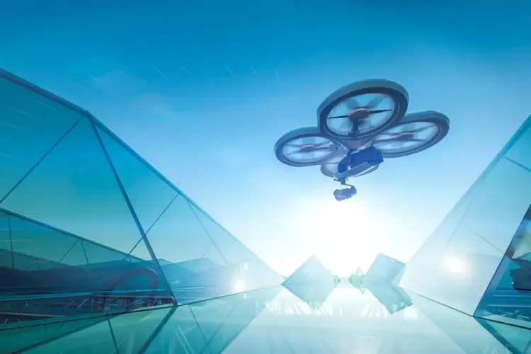 A camera drone with three propellers flies above a futuristic glass building under a bright blue sky.