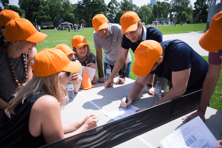 A group of people wearing orange caps gather around a picnic table outdoors, collaborating and writing on papers.