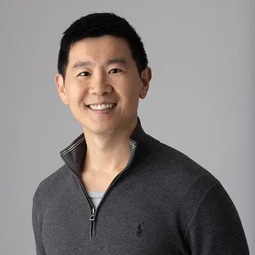 A smiling man with short black hair wearing a dark gray quarter-zip sweater over a light gray shirt stands in front of a light gray background.