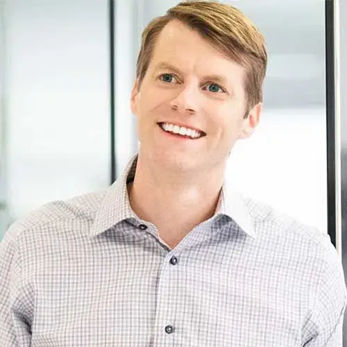 A smiling man with short light brown hair wearing a light-colored, checkered dress shirt stands indoors near a glass wall.